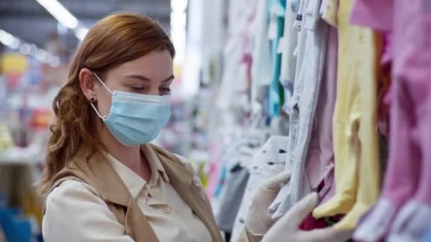 Karantänbutik, vårdande kvinna maskerad och handske på grund av pandemi och coronavirus väljer kläder för sitt barn — Stockvideo