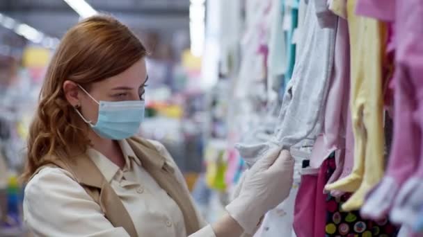 Pandemia y coronavirus, atractiva compradora con máscara y guantes para protegerse contra un virus mortal elige ropa de bebé en una tienda — Vídeo de stock