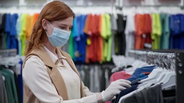 Compras pandémica, bastante joven comprador en máscara médica y guantes elige ropa nueva después de la cancelación de cuarentena — Vídeo de stock