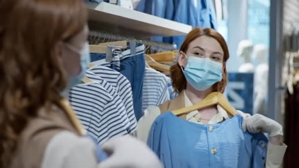 Koruyucu maskeli mutlu bir müşteri kızı portresi ve virüs ve enfeksiyondan korunmak için eldiven. Yeni bir elbise deniyorlar ve perakende giyim mağazasında aynaya bakıyorlar. — Stok video