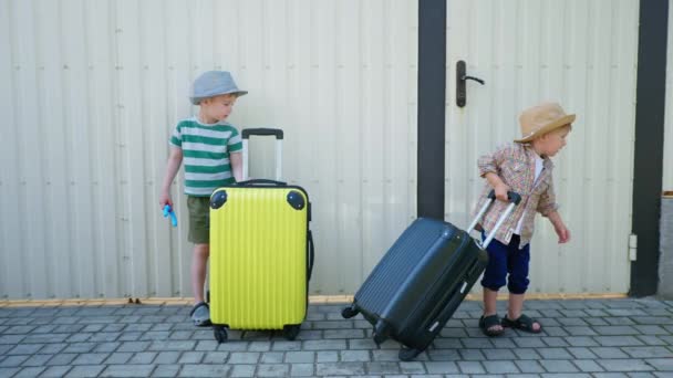 Viaje para los niños, los niños masculinos pequeños atractivos en sombreros del sol se divierten cerca de maletas durante las vacaciones de verano en el fondo de la cerca blanca — Vídeos de Stock