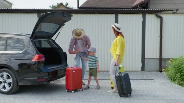 Весёлый муж и веселая жена с маленькими детьми-мужчинами кладут чемоданы в багажник для поездок с детьми на машине — стоковое видео
