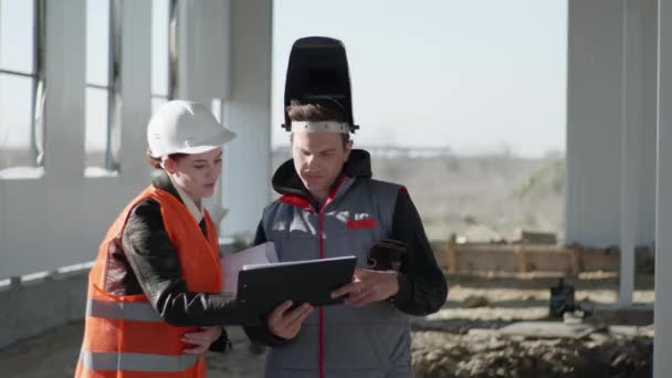 Engenheira feminina em capacete protetor e plantas em mãos em conjunto com um empregado discutir a execução do trabalho na construção de hangar — Vídeo de Stock