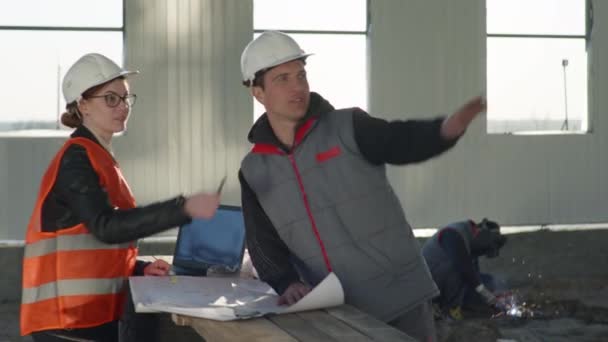 Чоловічі та жіночі інженери-будівельники в захисних жилетах і капелюхах перевіряють проект з офісних креслень на будівельному майданчику — стокове відео