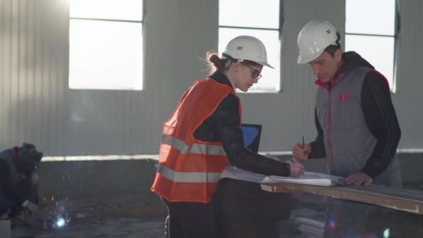 Byggnadsteknik, manliga och kvinnliga ingenjörer i skyddshjälmar diskutera projekt med hjälp av bärbara dator och ritningar — Stockvideo