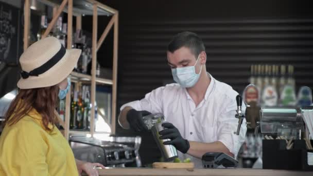 Vacanza quarantena, alla moda barista maschile in maschera medica versa ghiaccio e versa bere in vetro per preparare carino cocktail estivo ragazza in maschera — Video Stock