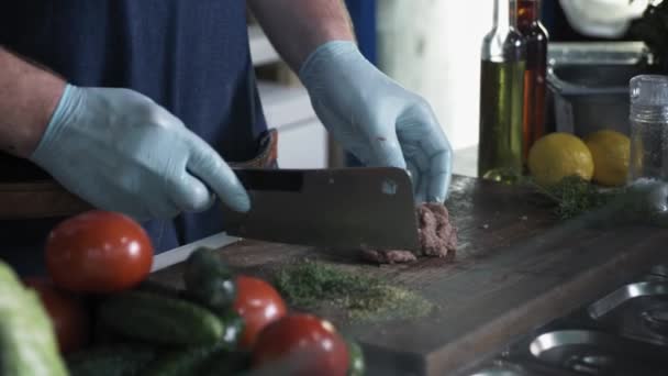 Επαγγελματίας αρσενικός μάγειρας κοπής μαχαίρι κρέατος για την παρασκευή burger σε ξύλο κοπής φόντο λαχανικών και μπαχαρικών, τα χέρια κοντά — Αρχείο Βίντεο