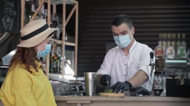 Karantänsemestrar, professionell bartender i medicinsk mask förbereder cocktail för kvinnliga besökare, man arbetar i baren lägger is och skakar skakapparat för att få dricka för flicka i skyddsmask — Stockvideo