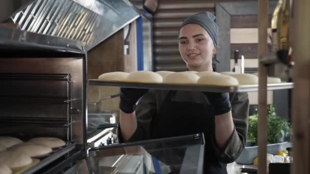 Productores locales, panadero femenino en uniforme pone bandeja para hornear con rebanadas de masa en el horno para obtener bollos frescos mientras se trabaja en la panadería — Vídeos de Stock