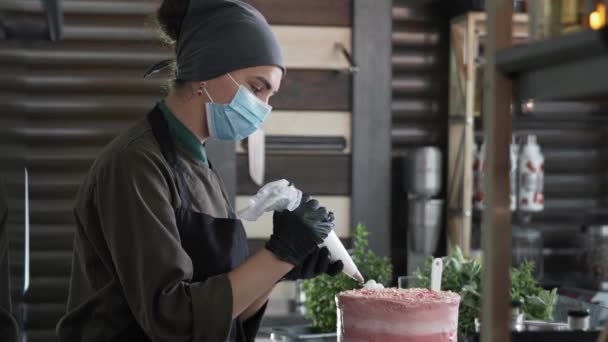 Kvinnlig bagare använder försiktighetsåtgärder i en medicinsk mask och handskar dekorerade hemmagjord söt kaka med grädde med hjälp av en bakelse väska ler och tittar på camer — Stockvideo