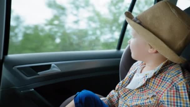 Estilo de vida, lindo niño varón en sombrero de paja mira por la ventana mientras está sentado en el asiento del coche, el niño se divierte y se alegra — Vídeos de Stock