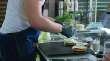Fast food, eldivenli şef hamburgerleri kesme tahtasının üzerinde pişirir. Lokantada ya da kafede çöreğe marul koyar.
