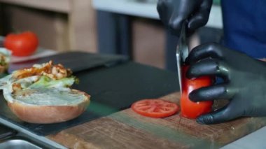 Yemek pişirmek, eldivenli aşçı kafeteryada çizburger yapmak için sulu domatesleri dilimleyip taze, kokulu soslu çöreğe, salataya ve kıyma ve peynire koyar.