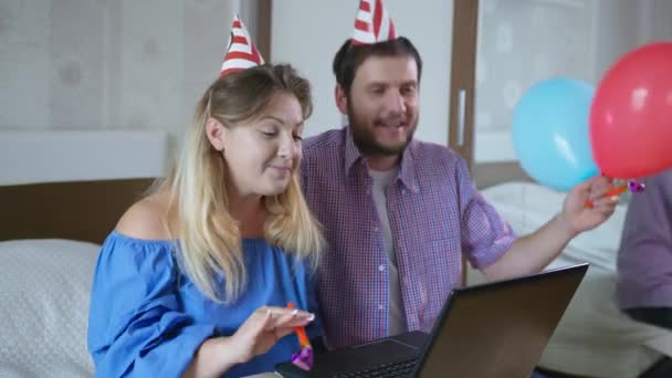 Fiesta en línea, joven pareja casada alegre crea un ambiente festivo con globos y gorras que soplan burbujas de aire y felicitando a su amigo de la familia en su cumpleaños por video que enlaza en el ordenador portátil mientras — Vídeo de stock