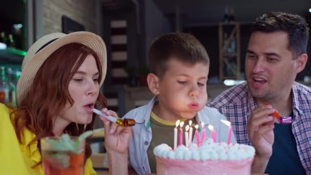 Веселі батьки з рогами святкують день народження зі своїм сином, маленький милий хлопчик розважається на святковій вечірці і вибухає свічками на торт, насолоджуючись весною в кафе або ресторані — стокове відео