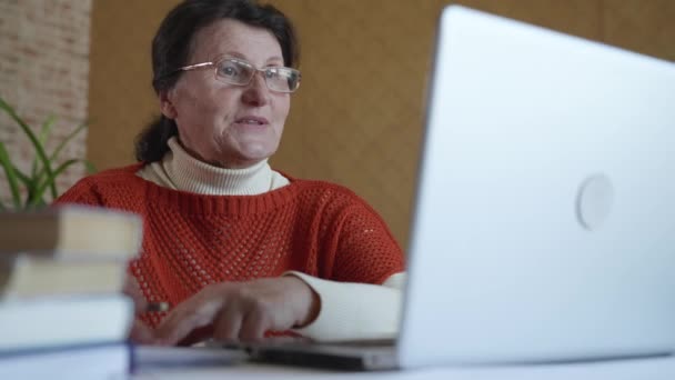 Σύγχρονες τεχνολογίες, χαρούμενη ηλικιωμένη γυναίκα με γυαλιά για την όραση μελετά την κατάρτιση σε απευθείας σύνδεση χρησιμοποιώντας το Διαδίκτυο και φορητό υπολογιστή κάθεται στο τραπέζι στο δωμάτιο — Αρχείο Βίντεο