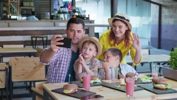 Festa de fast food, família alegre pais alegres amando mãe e pai com crianças do sexo masculino desfrutar de descanso no restaurante ou café comer hambúrgueres com batatas fritas e se comunicar com amigos por telefone — Vídeo de Stock