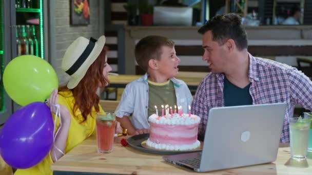 Pais alegres com seu filho amado celebram seu aniversário on-line e se comunicam com amigos usando uma câmera de laptop enquanto se sentam em uma mesa em um café Wi-Fi — Vídeo de Stock