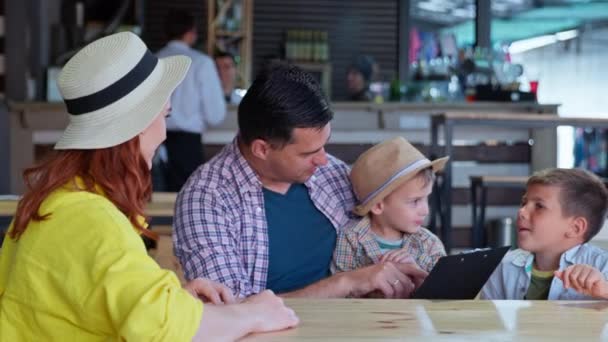Família feliz alegre de pais com crianças diverte-se à mesa no café e escolhe comida e bebidas do cardápio de restaurante — Vídeo de Stock