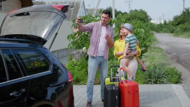 Radostné mužské a ženské rodiče se svým milovaným synem v náručí mluví prostřednictvím video spojení s přáteli v blízkosti kufříků pro rodinný výlet autem během — Stock video