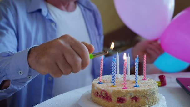 Святкування дня народження, літній чоловік з кульками в руках розважається створює святкову атмосферу і запалює святкові свічки на солодкому торті, крупним планом — стокове відео