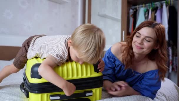 Sommerferie, lille søte guttebarn har det gøy med sin kjærlige mor og liker felles reisekofferter med ting og klær liggende på sofaen i rommet – stockvideo