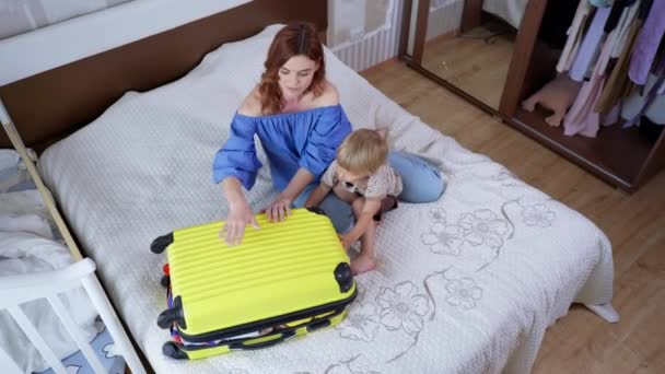 Viaje familiar, joven mamá alegre con su hijo varón trata de cerrar la maleta llena de ropa y pertenencias mientras se prepara para el viaje de verano al resort — Vídeo de stock
