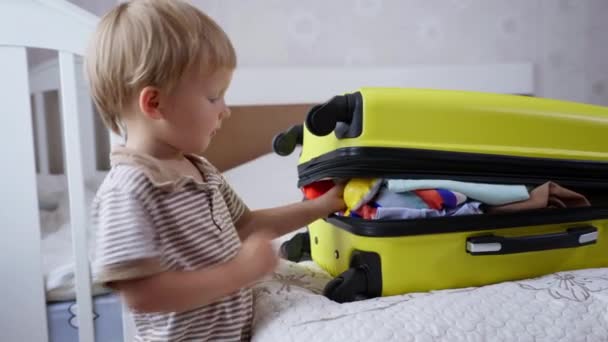 Vacaciones de verano, poco lindo niño masculino empaca para viaje familiar pone su juguete en la bolsa de viaje completa con ropa armarios cremallera — Vídeos de Stock