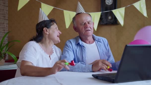 Вечірка онлайн, радісні бабусі і дідусі з повітряними кулями і трубами щасливі вітають своїх друзів або родину з днем народження на веб-камеру на ноутбуці, сидячи вдома за столом — стокове відео