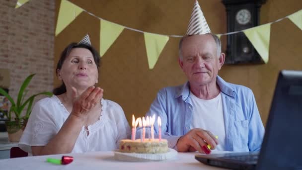 İzolasyon partisi, neşeli yaşlı kadın ve doğum günü pastası ve şapkalı adam mumlarla modern video iletişim teknolojisini laptopta kullan ve doğum gününü kendi kendine kutla — Stok video