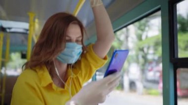 Sosyal mesafe, tıbbi maskeli ve eldivenli genç kız virüs ve kulaklıklarda enfeksiyondan korunmak için otobüs karantinası alanında seyahat ederken telefonda müzik dinliyor.