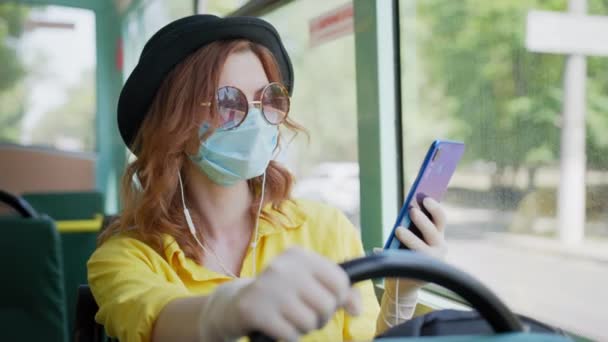 Junge Frau mit Hut mit Brille und Kopfhörer hört Musik, während sie in öffentlichen Verkehrsmitteln unterwegs ist und trifft Sicherheitsvorkehrungen trägt medizinische Schutzmaske und Handschuhe, um sich vor dem Virus zu schützen — Stockvideo