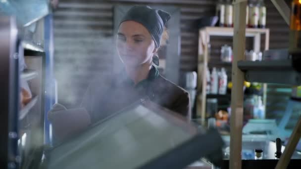 Pequeña empresa, panadero femenino en guantes y uniforme saca pan recién horneado del horno caliente, pastelera abre un horno y da bandeja para hornear con hornear y fumar, cocinar — Vídeos de Stock