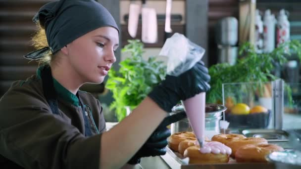 Sobremesa, confeiteiro chef bonito em luvas, espreme creme de proteína grossa de saco de pastelaria para decorar deliciosos donuts frescos, comida insalubre — Vídeo de Stock