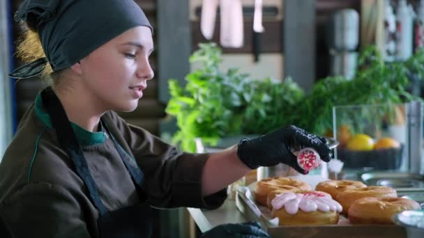 Charmig konditor flicka som arbetar i köket i bageri och dekorera söta stekta munkar med handgjort pulver, kvinnlig kock ler och tittar på kameran — Stockvideo
