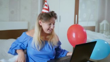 Sosyal mesafeli, elinde balon ve piposu olan evcil kedisi olan genç bir kadın, karantina ve karantina yüzünden evinde yatarken bilgisayardan arkadaşlarını tebrik ediyor.