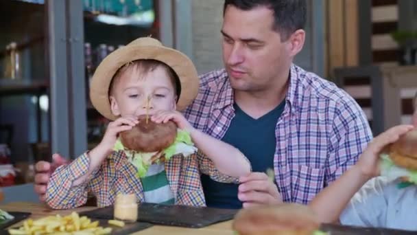 Fast food, malý atraktivní chlapec se slamákem jí ústa zalévání šťavnatý burger sedí radostný táta v náručí při relaxaci v kavárně — Stock video