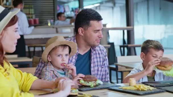 Mand og kvinde med mandlige børn nyder at spise velsmagende saftige burgere med pommes frites sidder ved bordet i cafe – Stock-video