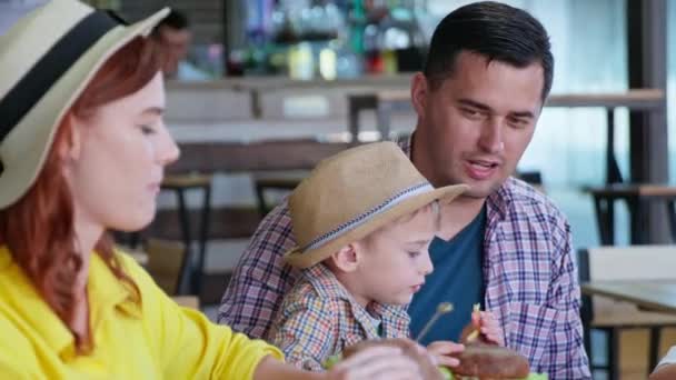 매력적 인 부모는 귀여운 남아와 함께 가족이 휴가를 보내는 식당에서 맛있는 햄버거와 감자튀김을 즐긴다 — 비디오