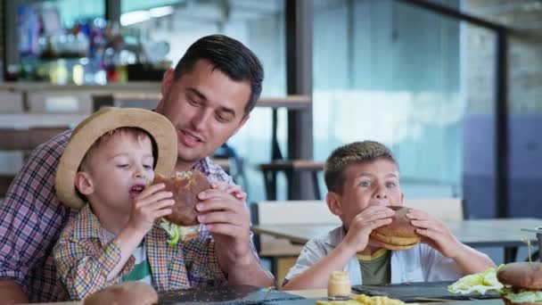 Kinder essen, fröhliche männliche Eltern genießen zusammen mit ihren hungrigen Söhnen leckere Burger — Stockvideo