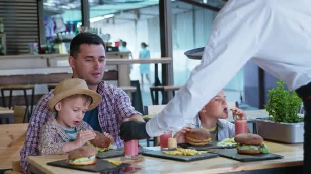 Pai carinhoso com seus filhos masculinos amados se divertir no café popular e come batatas fritas à mesa, enquanto garçom traz delicioso suco de frutas, pequenos negócios — Vídeo de Stock