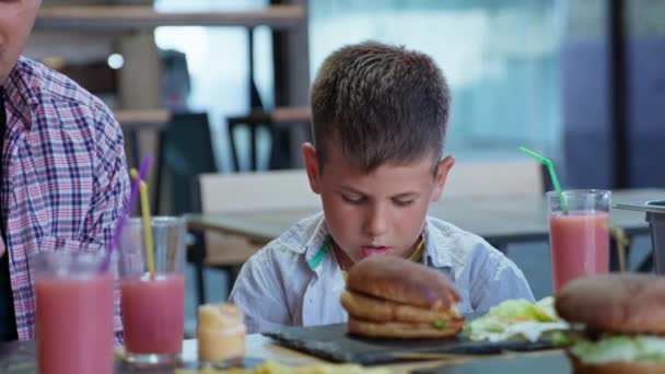 Fast food, hongerige mannelijke kinderen met hun vader eten smakelijke sappige vleesburgers met frietjes en vruchtensap tijdens een familiediner in café — Stockvideo