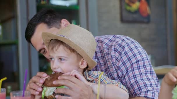 Podvýživa, malý roztomilý chlapec jí rychlé občerstvení vydatné maso burger se svým milujícím otcem během rodinné dovolené v kavárně — Stock video
