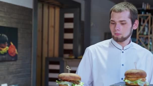 Pěkný číšník dělá chutné šťavnaté burgery radostné a hladové návštěvníky šťastné rodiny s dětmi baví v kavárně v kavárně — Stock video