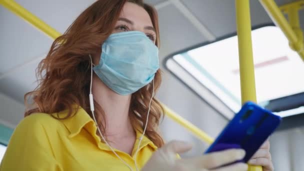 Портрет веселої дівчини в громадському транспорті з рукавичками носить медичну маску для захисту від вірусів та інфекцій і слухає музику з навушниками на смартфоні під час подорожі на автобусі після — стокове відео