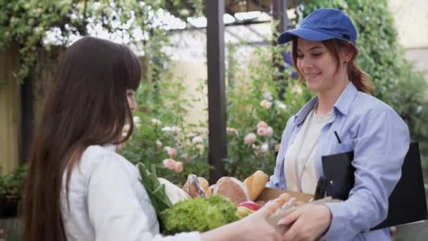 Yemek teslimatı, neşeli kadın kurye evdeki bir çevrimiçi dükkandan sipariş edilen genç bayana bir kutu yiyecek getiriyor. — Stok video