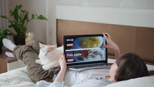 Delivery restaurant, girl uses her laptop to order ready-made meals online using modern customer service during the movie and pandemic, woman lying on couch buying groceries online ordering fast food — Stock Video