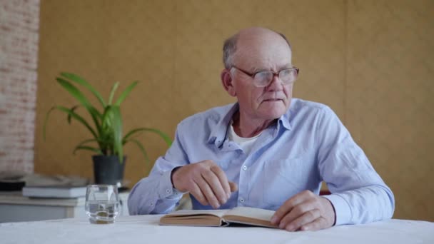 Attractive elderly male pensioner with glasses for vision drinks clean cool water from transparent glass during interesting book sitting at table in room — Stock Video