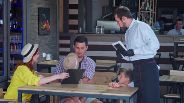 Mann und Frau mit ihren fröhlichen Söhnen genießen es, Zeit miteinander zu verbringen und Kellner zu bestellen, während sie sich im Café entspannen — Stockvideo