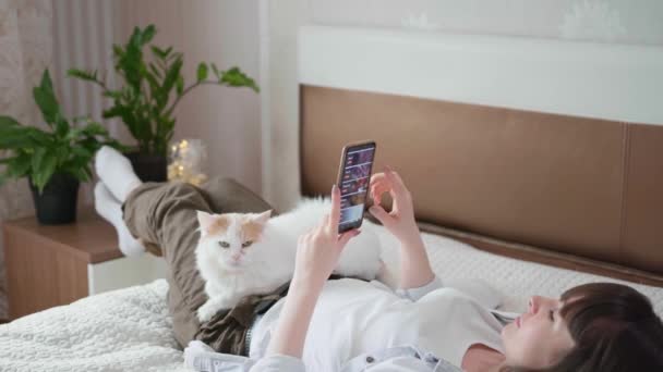 Mujer joven utiliza teléfono inteligente y ordena comida en línea utilizando Internet mientras está acostado en casa en la cama con el gato durante el auto-aislamiento — Vídeos de Stock